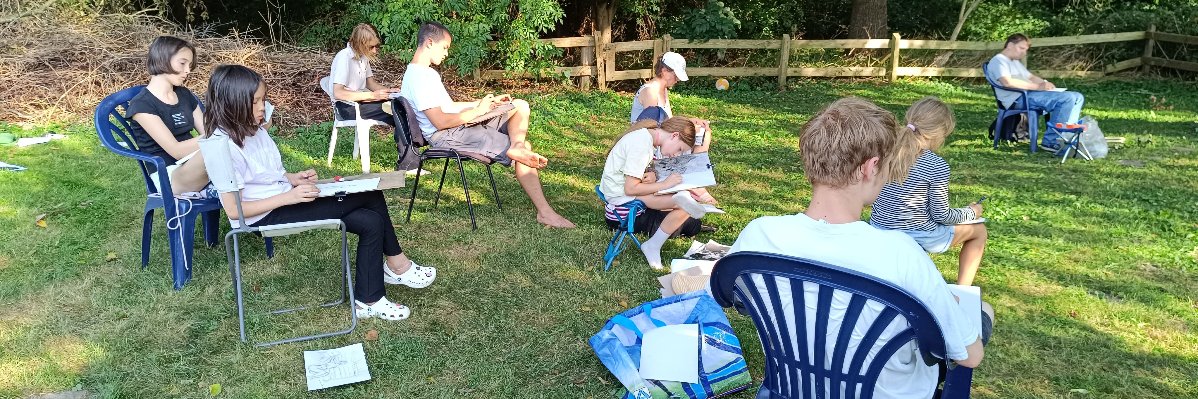 Gruppe von Kindern und Jugendlichen zeichnet im Freien auf einer Wiese, umgeben von Bäumen und einem Holzzaun.