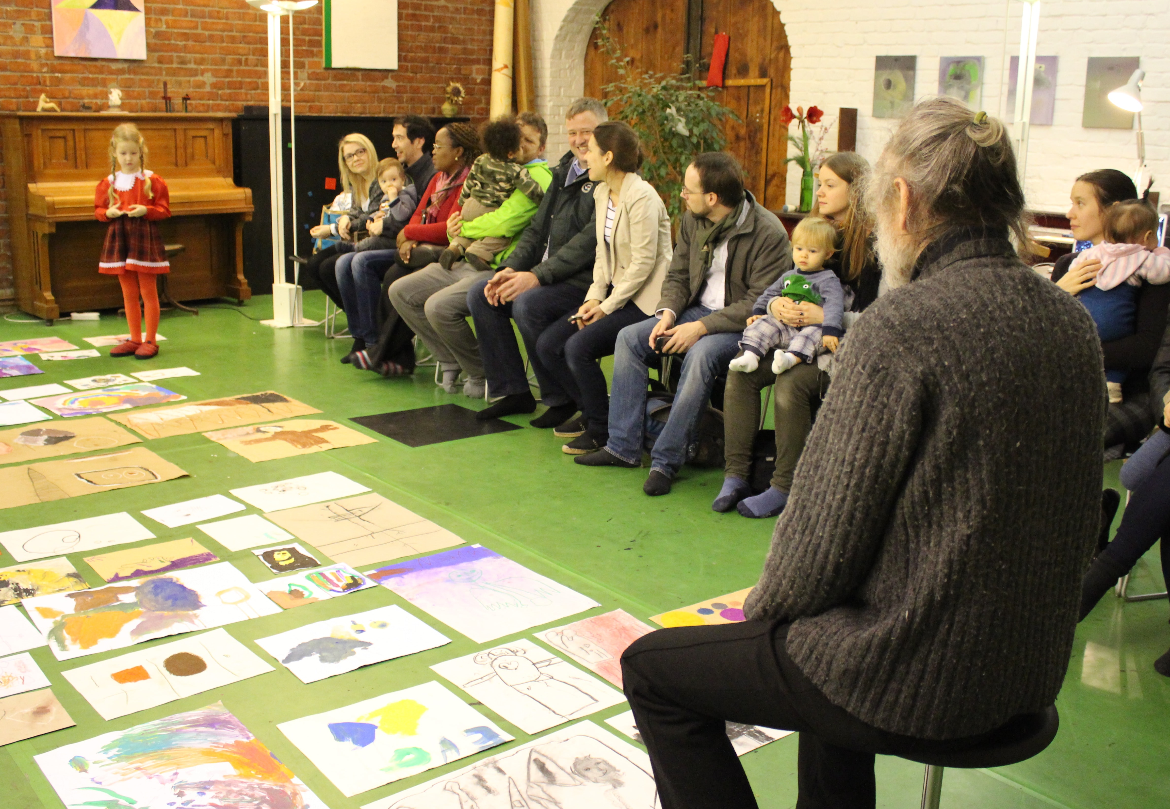 Eine junge Künstlerin präsentiert ihre Werke vor einem Publikum, während zahlreiche Kinderbilder auf dem Boden ausgelegt sind.